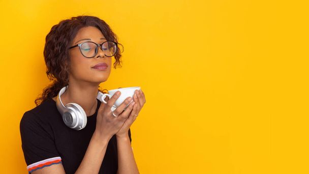 African-american beautiful young womans portrait with wireless headphones on yellow studio background, emotional and expressive. Copyspace for ad. - Fotó, kép
