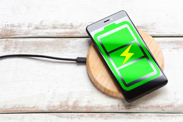 現代技術の概念。ワイヤレス充電器で緑のバッテリーアイコンと黒のスマートフォンです。水平方向の縞模様の白い木製の背景。コピースペース。平線. - 写真・画像