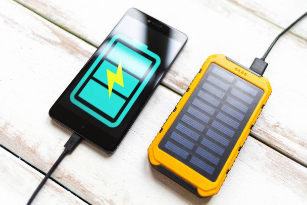 Fogalom a modern technológiák és a hatalom bank. Okostelefon egy türkiz akkumulátor ikon, töltött egy hordozható napelemes töltő. Fehér fa háttér csíkos. Közelről.. - Fotó, kép