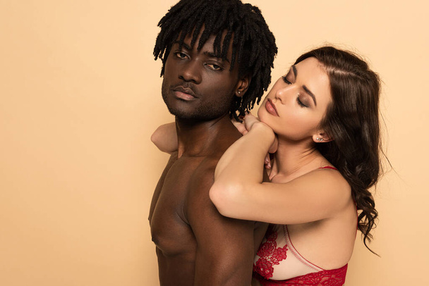 sexy vriendin knuffelen shirtless afrikaans amerikaans vriend geïsoleerd op beige - Foto, afbeelding