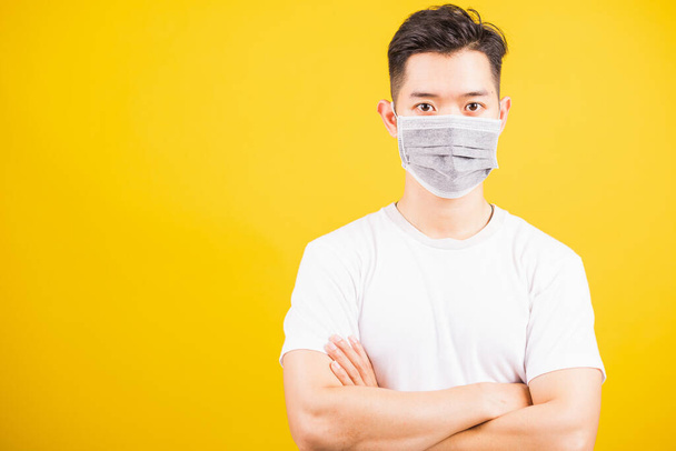Closeup Asian young handsome man wearing face mask protective germ virus and stand crossed arm, studio shot isolated on yellow background and copy space, medical outbreak coronavirus COVID-19 concept - Фото, изображение