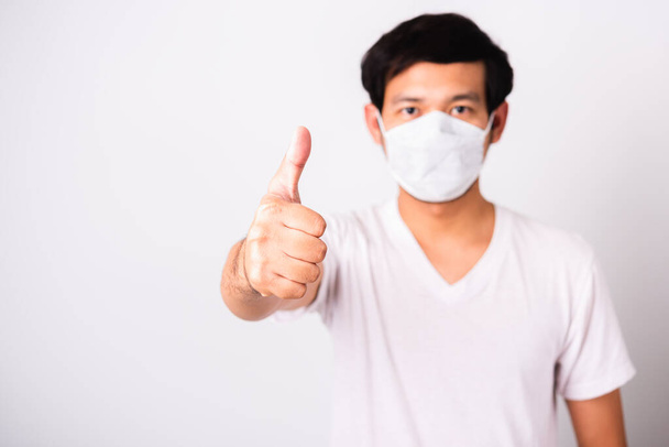 Asiatischer gutaussehender Mann mit chirurgischem hygienischen Schutztuch Gesichtsmaske gegen Coronavirus zeigt er Daumen für gut, Studioaufnahme isolierten weißen Hintergrund, COVID-19 medizinisches Konzept - Foto, Bild