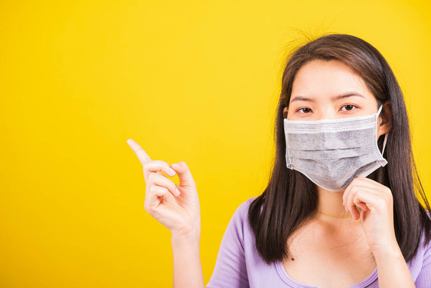 Asian young teen woman wearing face mask protective against coronavirus or COVID-19 virus or filter dust, air pollution her point finger to side space, studio shot isolated yellow background - Foto, Imagem