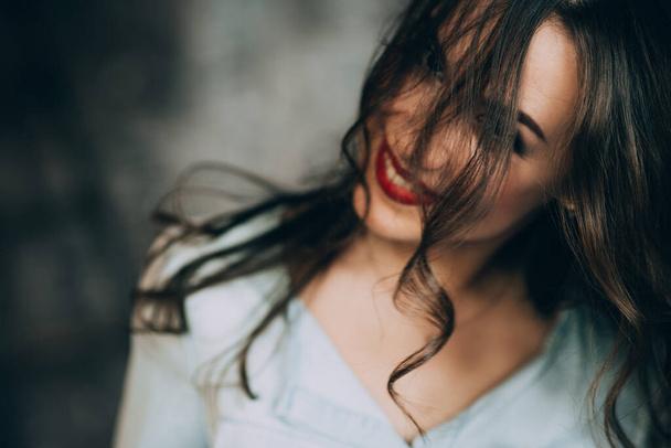 Όμορφη νεαρή γυναίκα με τέλεια μαύρα σγουρά μαλλιά και όμορφο make-up με παχουλά κόκκινα χείλη σε ένα τζιν πουκάμισο κοιτάζει την κάμερα μέσα από τα μαλλιά της και παιχνιδιάρικα χαμόγελα. Μαλακή επιλεκτική εστίαση. - Φωτογραφία, εικόνα