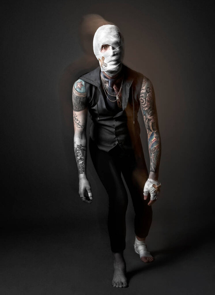 Τραυματισμένος, παράξενος άντρας με μαύρο γιλέκο και παντελόνι, τα χέρια του καλυμμένα με τατουάζ, το κεφάλι, το χέρι και το πόδι του τυλιγμένα με επιδέσμους.. - Φωτογραφία, εικόνα