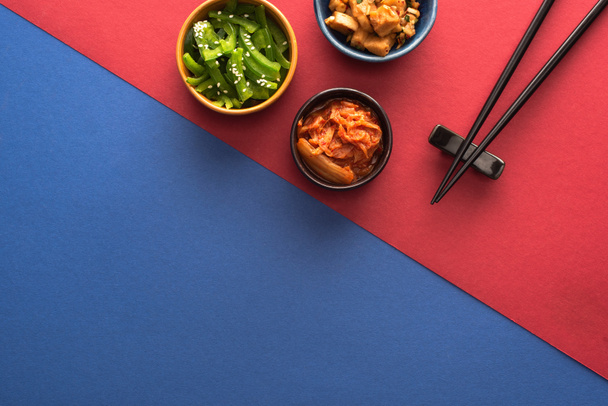 top view of bowls with spicy korean food near chopsticks on blue and crimson   - Фото, изображение