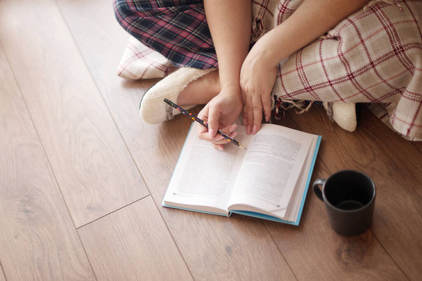 Ruce dívky v domácích šatech, čte knihu, sedí na dřevěné podlaze s tužkou v ruce, hrnek na podlaze. koncepce domácích úkolů, domácí odpočinek - Fotografie, Obrázek