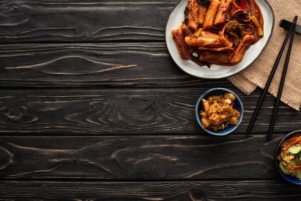 vista superior de topokki coreano picante perto de batatas assadas, pepinos em conserva, pauzinhos e guardanapo de algodão na superfície de madeira
  - Foto, Imagem