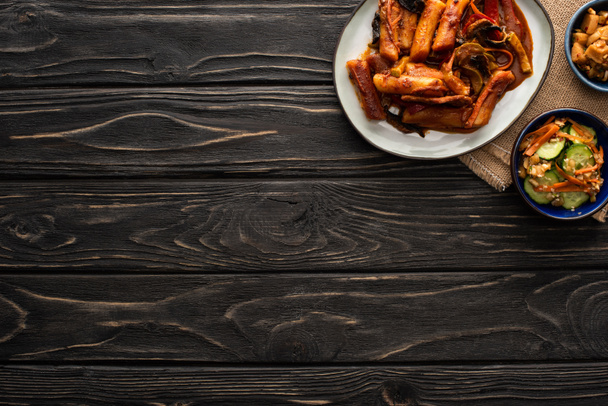 vista superior de topokki picante perto de batatas refogadas coreanas, pepinos em conserva, pauzinhos e guardanapo de algodão na superfície de madeira
  - Foto, Imagem