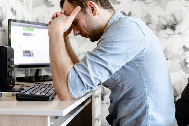 νεαρός άνδρας ελεύθερος επαγγελματίας κάθεται στο σπίτι σε έναν υπολογιστή και έσκυψε το κεφάλι του στην απογοήτευση - Φωτογραφία, εικόνα