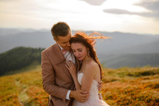 The bride and groom hug and tenderly with each other. Sunset. Wedding photo on a background of autumn mountains. A strong wind inflates hair and dress. Close-up. - Foto, Bild