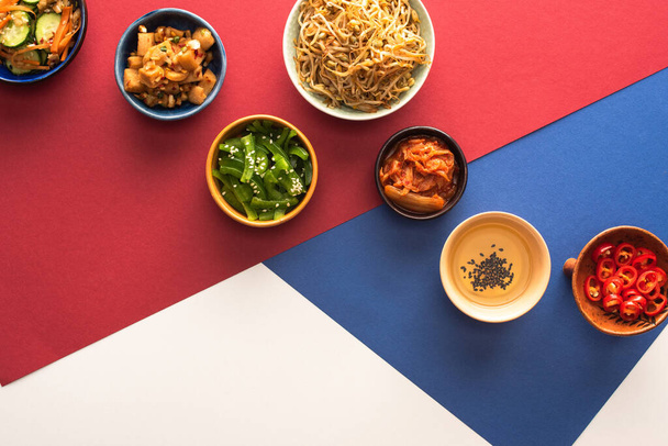Draufsicht auf Schalen mit traditionellen koreanischen Beilagen und Öl auf blau, purpurrot und weiß  - Foto, Bild