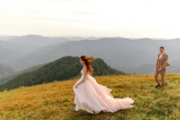 The bride and groom go next to each other. Sunset. Wedding photo on a background of autumn mountains. A strong wind inflates hair and dress. - Фото, изображение