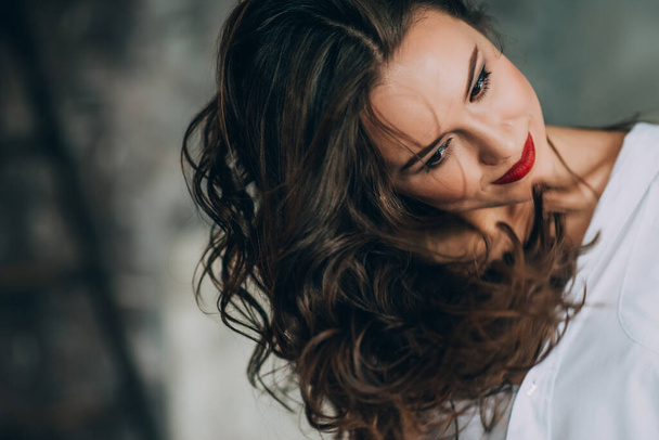 Schöne junge Frau mit perfekt dunkelgelockten Haaren und wunderschönem Make-up mit pausbäckigen roten Lippen im weißen Hemd. Weiche selektive Fokussierung. - Foto, Bild