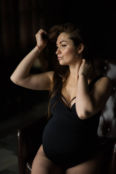 Pregnant women, photoshoot in dark key sit on the armchair - Photo, Image