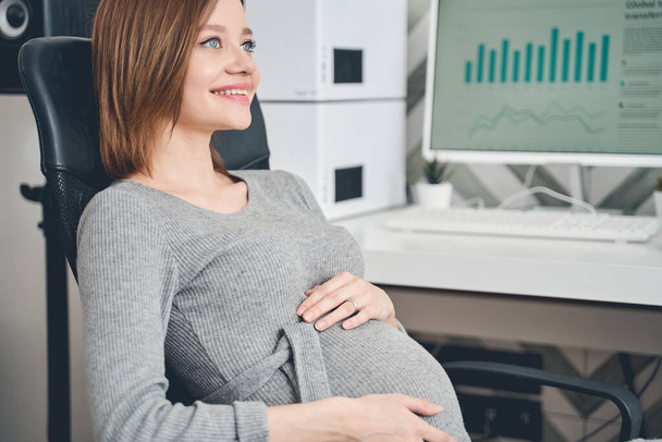 Cheerful pregnant lady sitting in office chair at work - Photo, image