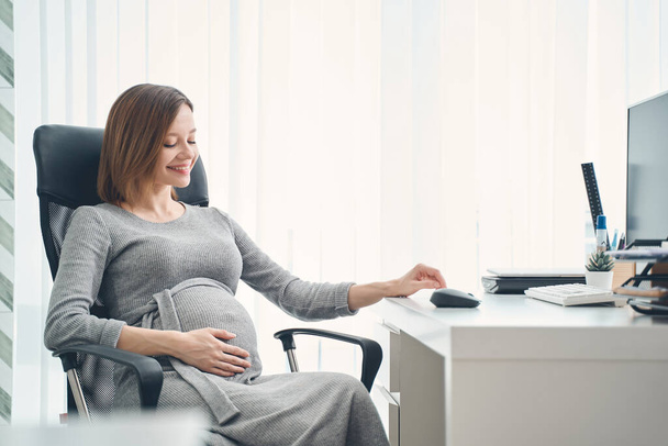Cute expectant woman sitting at office desk and smiling - Foto, Imagen