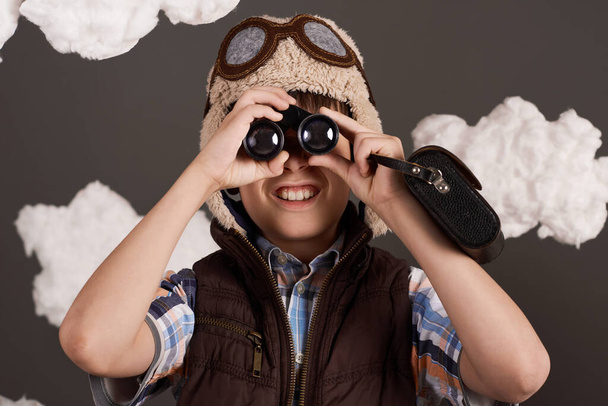 um menino brinca com binóculos e sonhos de se tornar um piloto, vestido com uma jaqueta estilo retro e capacete com óculos, nuvens de lã de algodão, fundo cinza, matizado em marrom
 - Foto, Imagem