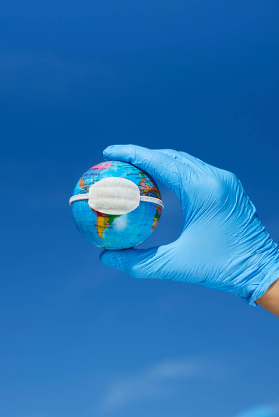 close-up van een man, met blauwe chirurgische handschoenen, met een aardbol met een beschermend masker, waarop de wereldwijde coronaviruspandemie of de luchtverontreiniging in de planeet is afgebeeld - Foto, afbeelding