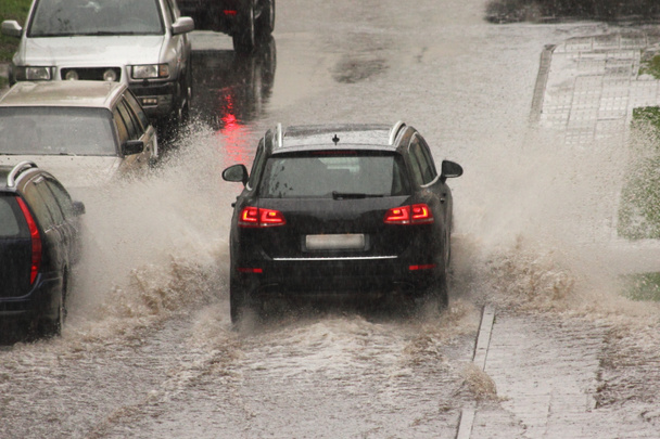 Siyah bir Alman lüks SUV aracı, bir bahar günü, arka planda sağanak yağmurda, park halindeki arabaların yanında, sokağın aşağısına doğru ağır bir sprey püskürtüyle hızla gider. - Fotoğraf, Görsel