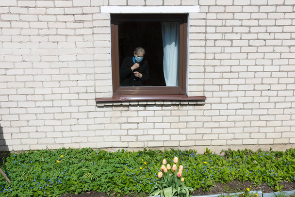 Μια γιαγιά με προστατευτική μάσκα κοιτάει έξω από το παράθυρο την αυτο-απομόνωση.. - Φωτογραφία, εικόνα