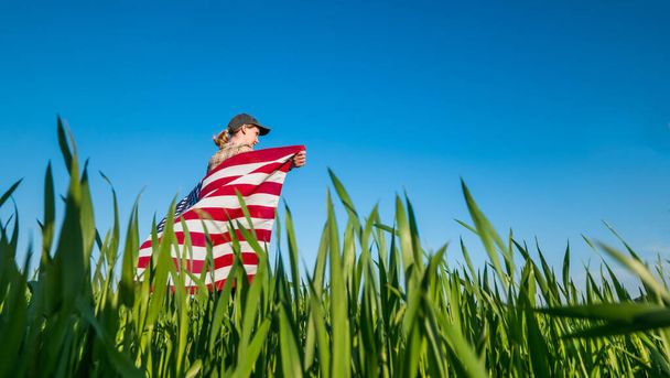 A farmer with a U.S. flag on his shoulders stands in a field of green wheat - Fotografie, Obrázek