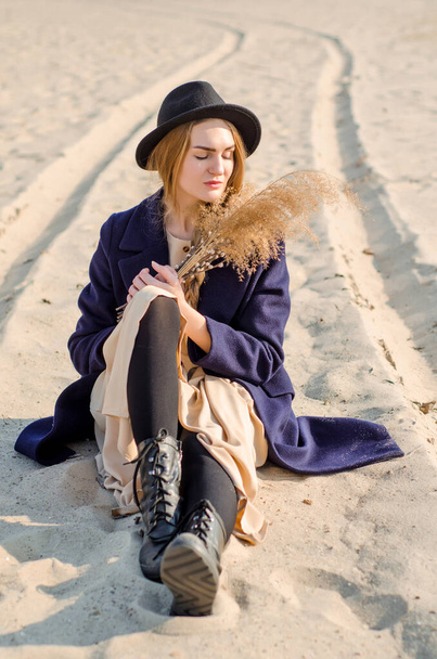 Young caucasian blonde girl in dress, coat, hat on beach, under rays of sun with closed eyes, dream about something - Foto, imagen