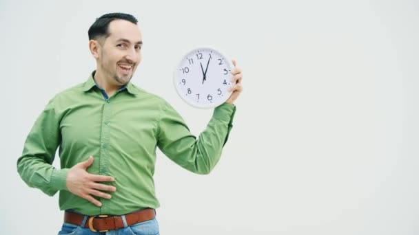 Funny young man in casual clothes with the clock in hand, patting, holding and blowing his belly making grimaces. Expressive facial expression. - Záběry, video