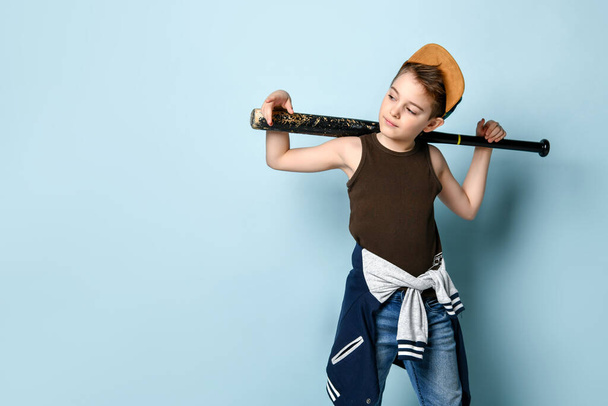 Sly little schoolboy stands near the wall looking aside holding baseball bat on shoulders. Three quarter length portrait isolated on blue background - 写真・画像