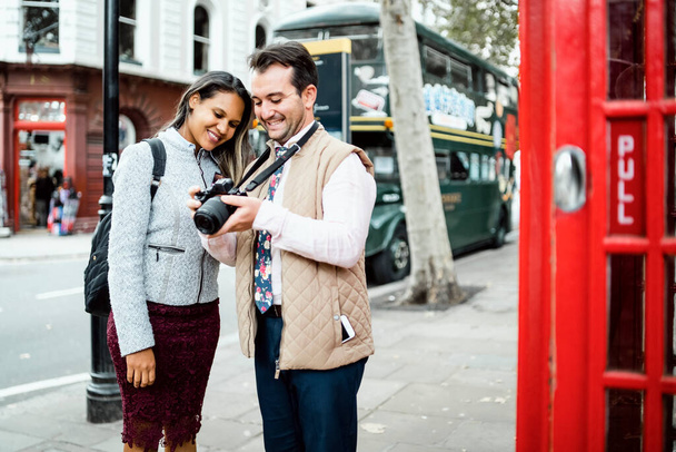 Ευτυχισμένο ζευγάρι που ταξιδεύουν κριτική φωτογραφίες στην κάμερα τους στο Λονδίνο, Ηνωμένο Βασίλειο. Διπλό υπερυψωμένο λεωφορείο και κόκκινο τηλεφωνικό θάλαμο σε πλαίσιο - Φωτογραφία, εικόνα