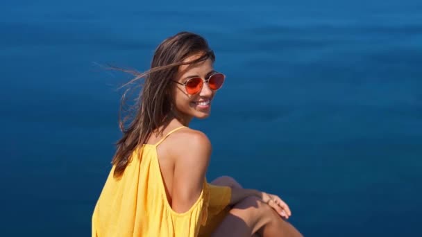 szczęśliwa kobieta w jasnożółtej sukience i czerwonych okularach siedzi na krawędzi klifu przed niesamowite krajobraz morski. - Materiał filmowy, wideo