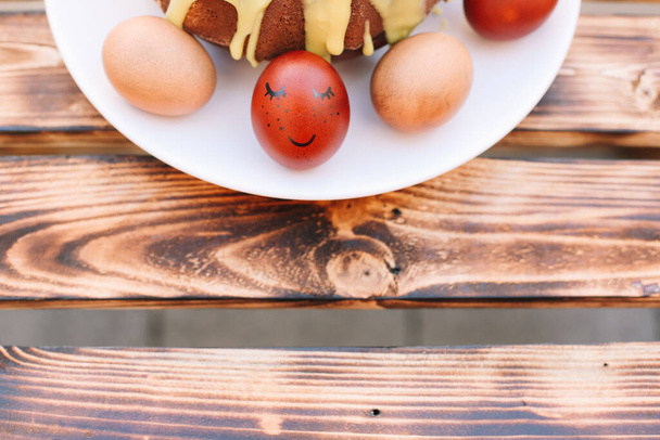 Huevo de Pascua rojo con una sonrisa alrededor de los huevos y pastel de Pascua en el plato blanco en el fondo de madera
 - Foto, Imagen