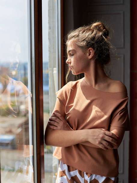 Young brunette woman, wearing beige jersey pajamas, standing neat big window in dark room in the morning. Hen party celebration. Fashion photography. Bridesmaid on wedding morning. - Fotografie, Obrázek
