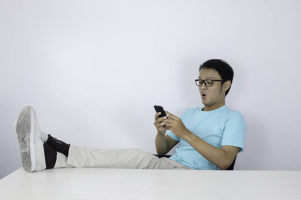 Fiatal ázsiai férfi boldog arccal azzal, amit egy mobiltelefonon lát lábakkal az asztalon. Indonéz férfi kék ingben.. - Fotó, kép