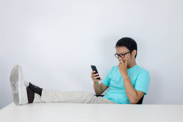 Young Asian Man with happy face with what he see on a mobile phone with leg on the table. Indonesian man wearing blue shirt. - Photo, Image