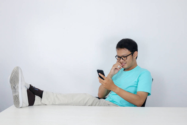 Young Asian Man with happy face with what he see on a mobile phone with leg on the table. Indonesian man wearing blue shirt. - Φωτογραφία, εικόνα