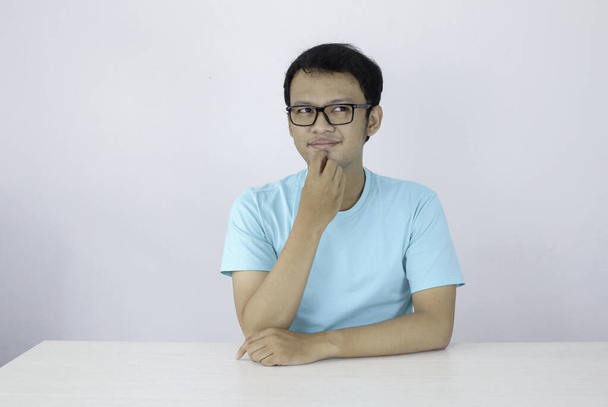 Young Asian man wear blue shirt and glasses with thinking and looking idea gesture in above empty space - Fotó, kép