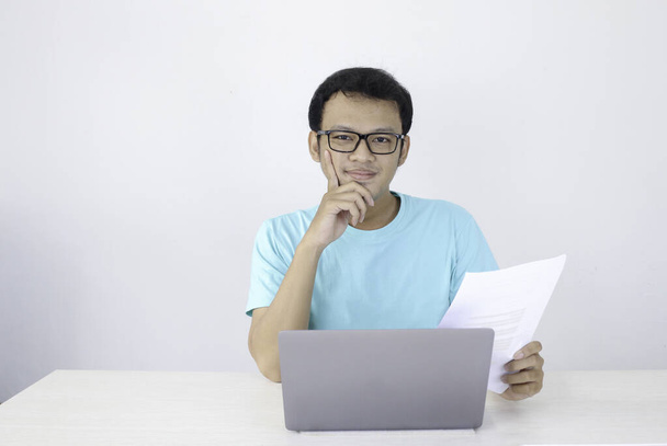 Młody Azjata jest uśmiechnięty i szczęśliwy, gdy pracuje nad laptopem i dokumentem. Indonezyjczyk w niebieskiej koszuli. - Zdjęcie, obraz