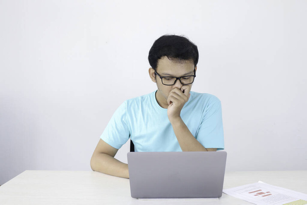 Young Asian Man is serious and focus when working on a laptop and document on the table. Indonesian man wearing blue shirt. - Фото, изображение