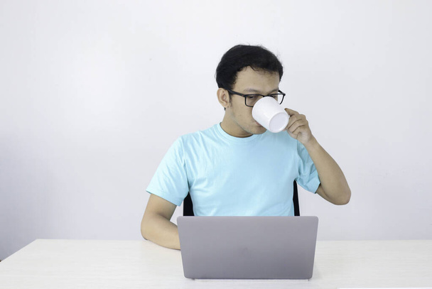Joven asiático hombre es serio cuando el trabajo en el ordenador portátil con beber café en vidrio blanco. Indonesia Hombre usar camisa azul Fondo gris aislado
. - Foto, imagen