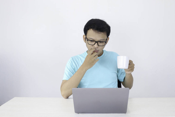 Молодой азиат серьезен, когда работает в ноутбуке с кофе в белом стакане. Индонезия Мужчина носить голубую рубашку Изолированный серый фон
. - Фото, изображение