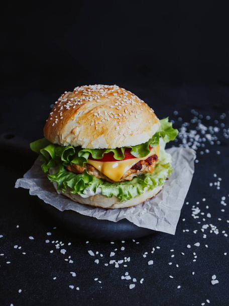 Appetizing fresh homemade burger, with chicken cutlet, lettuce, tomatoes, cheese and sauce. On a wooden board on a black background - Фото, изображение