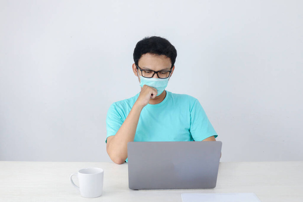 Молодий азіатський чоловік у медичній масці є серйозним і зосередженим на роботі над ноутбуком на столі. Індонезійський чоловік у синій сорочці.. - Фото, зображення