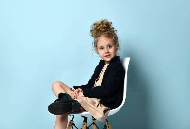 Beautiful little blonde girl in beige dress and black jacket sitting cross-legged in office chair with confident smile, isolated on blue - Fotoğraf, Görsel
