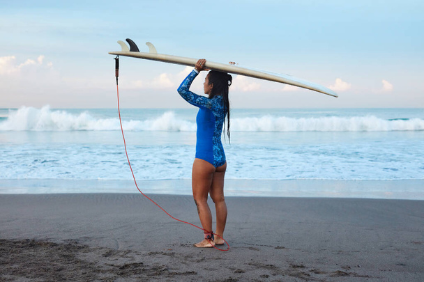 Chica Surfista. Mujer surfeando con tabla de surf en la cabeza de pie en Sandy Beach. Morena en traje azul yendo a surfear en el mar. Deporte acuático para un estilo de vida activo
. - Foto, Imagen