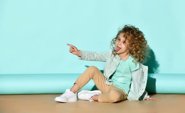 Little blonde curly lady in casual clothes. Showing tongue, pointing at something, sitting on floor over turquoise background - Photo, Image