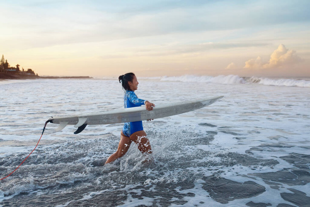 Surf In Ocean. Surfing Girl With Surfboard Walking Into Ocean. Young Surfer In Blue Wetsuit Going To Swim In Sea At Sunset. Water Sport Active Lifestyle.  - 写真・画像