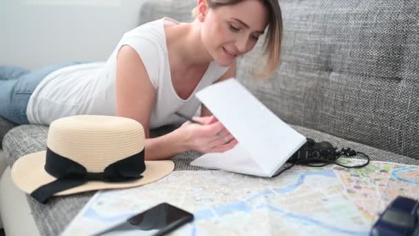 Νεαρή ευτυχισμένη γυναίκα σχεδιάζει ταξίδι διακοπών στο σπίτι μετά το τέλος της πανδημίας και της καραντίνας. Κορίτσι κοιτάζοντας ένα χάρτη γράφει πληροφορίες σε ένα σημειωματάριο.  - Πλάνα, βίντεο