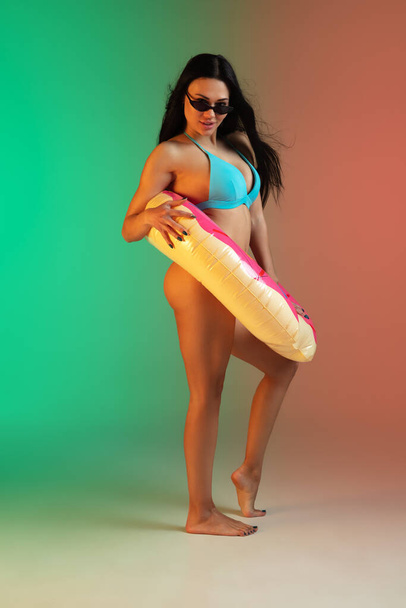 Fashion portrait of young fit and sportive woman in blue luxury swimwear with rubber donut and stylish sunglasses on gradient background. Perfect body ready for summertime. - Zdjęcie, obraz