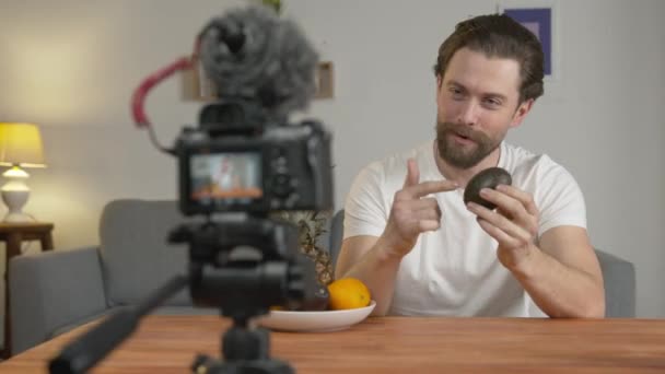 Mladý muž, vlogger točí video, sedí u stolu před kamerou a mluví o ovoci a jejich výhodách, avokádo - Záběry, video